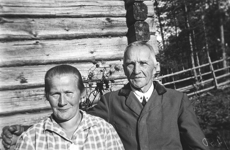 Mustavalkoinen kuva miehestä ja naisesta hirsitalon edessä.